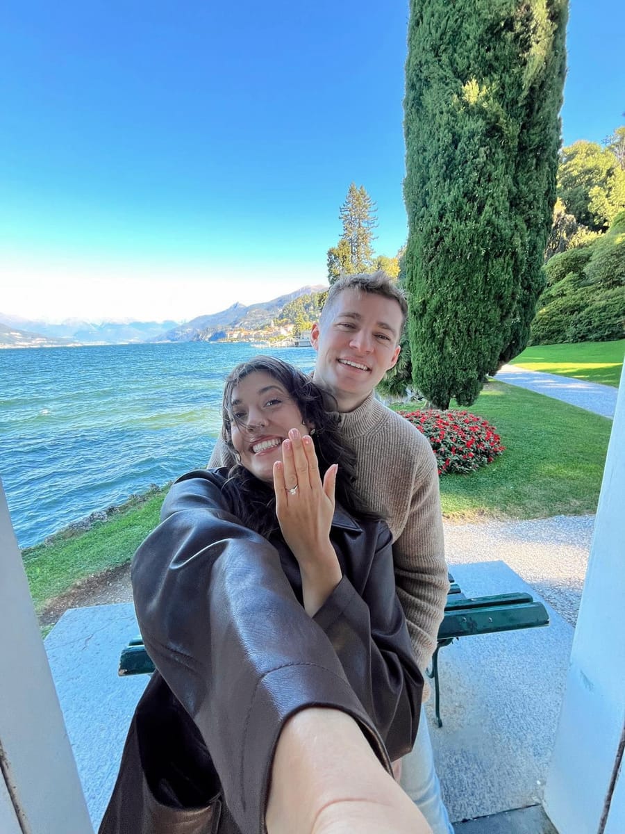 Justyna showing her engagement ring, with Lewis behind her, beside a lake